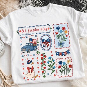 May include: White t-shirt with patriotic illustrations. Features a blue truck with an American flag, floral bouquets, a goose in a patriotic hat, and the text "let freedom ring". The design is in red, white, and blue.