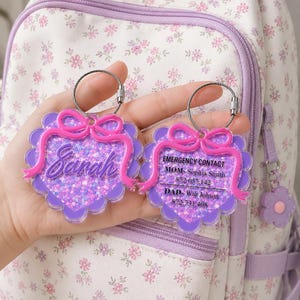 May include: Two heart-shaped acrylic bag tags with purple glitter centers and scalloped edges. One tag features the name Sarah in raised pink script, while the other displays emergency contact information for Mom and Dad with phone numbers.