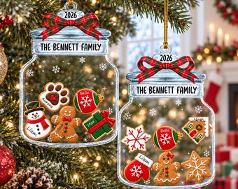 May include: Two clear glass jar ornaments featuring the text THE BENNETT FAMILY and 2026, topped with red and green plaid bows. Inside the jars are various gingerbread-style cookies shaped like a paw, snowman, gift, mitten, and star.
