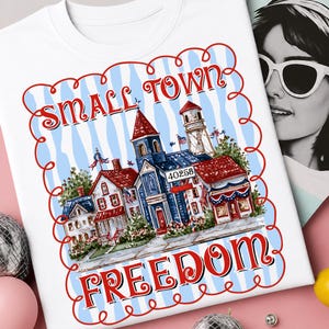 Könnte beinhalten: Weißes T-Shirt mit einer farbenfrohen Illustration einer Kleinstadt mit roten, weißen und blauen Gebäuden. Die Wörter "SMALL TOWN" und "FREEDOM" sind in roter Schrift geschrieben. Das Design wird von einem roten Rand umrahmt.