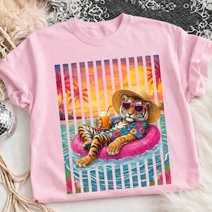 May include: Pink t-shirt featuring a tiger wearing sunglasses, a straw hat, and a Hawaiian shirt, relaxing on a pink inflatable ring in a pool. The tiger is holding a drink with a straw. The background shows a sunset with palm trees.