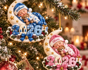 May include: Two Christmas ornaments shaped like crescent moons, each featuring a sleeping baby in pajamas holding a teddy bear. One ornament is blue with the name William, the other is pink with the name Olivia. Both display the year 2026.