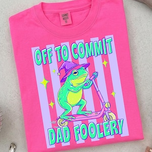 May include: A bright pink t-shirt with the text "OFF TO COMMIT DAD FOOLERY". The graphic features a green frog wearing a purple wizard hat and riding a pink scooter. The shirt has a crew neck and a small tag on the upper left.
