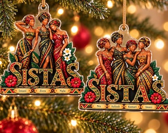 May include: Two clear acrylic Christmas ornaments featuring illustrations of women in patterned dresses and headwraps. The left ornament displays the text SISTAS with names Imani, Zora, and Nyla. The right ornament displays SISTAS with Nia, Aaliyah, Kamari, and Imani.