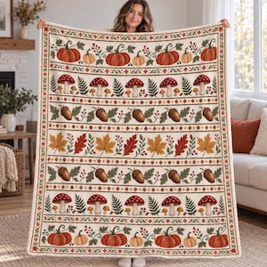 May include: A cream-coloured throw blanket featuring a repeating horizontal pattern of orange pumpkins, red and white mushrooms, brown acorns, and multicoloured autumn leaves, all framed by a decorative border with small red berries and green foliage.