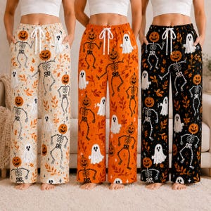 May include: Three pairs of wide-leg lounge pants featuring a Halloween pattern of skeletons with pumpkin heads, white ghosts, and orange leaves. The pants are available in cream, orange, and black, each with a white drawstring waist tie.