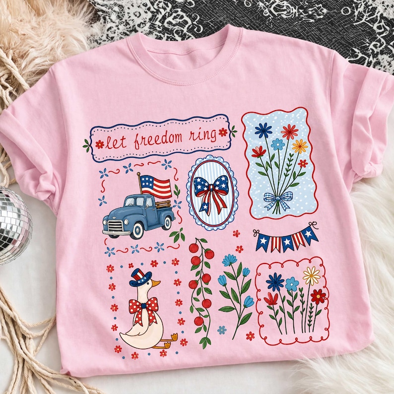 May include: Pale pink t-shirt with patriotic designs. Includes a blue truck with an American flag, a floral bouquet, and a goose in a hat and bow tie. The words "let freedom ring" are printed at the top.