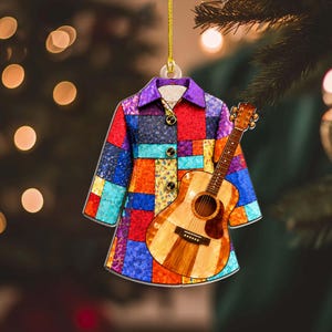 May include: A flat, hanging ornament shaped like a long-sleeved coat with a vibrant, multi-coloured patchwork pattern in shades of red, blue, orange, and purple, featuring a light-coloured acoustic guitar layered over the front of the garment.