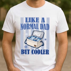 May include: White t-shirt with a blue graphic. The graphic reads "LIKE A NORMAL DAD BUT COOLER" and features a cooler filled with cans and ice. Blue stripes and snowflakes are also part of the design.
