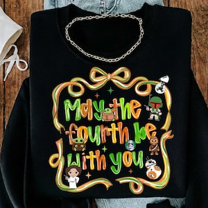 May include: Black sweatshirt with the phrase "May the fourth be with you" in green and orange lettering. The design includes cartoon characters from the Star Wars franchise, such as Baby Yoda, Boba Fett, and Princess Leia.