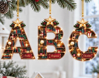 May include: Three hanging ornaments shaped like the letters A, B, and C. Each letter features a miniature bookshelf interior with tiny books, a globe, a deer, a Christmas tree, and a gold star topper. The names Amelia, Benjamin, and Charlotte are printed on red plaques.