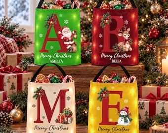 May include: Four cylindrical fabric Christmas gift baskets with black handles and integrated LED lights. Each basket features a large letter, a holiday graphic, the text Merry Christmas, and a personalized name: Amelia, Bella, Mia, or Emma.