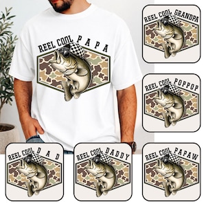 May include: White t-shirt with a "REEL COOL PAPA" graphic. The graphic features a cartoon bass wearing sunglasses and a checkered hat, set against a camouflage background. Additional designs include "GRANDPA", "POPOOP", "DAD", "DADDY", and "PAPAW".