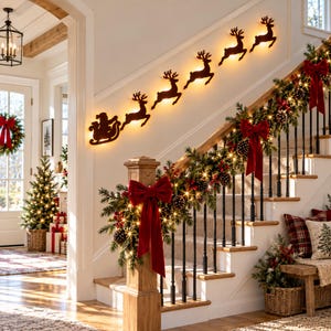 Puede incluir: Una escalera decorada con una guirnalda verde con piñas, bayas rojas y grandes lazos de terciopelo rojo. En la pared sobre la escalera, una serie de siluetas oscuras de Santa en un trineo y seis renos están iluminadas por una luz cálida.