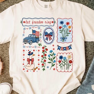 May include: Cream-coloured t-shirt with patriotic graphics. Designs include a blue truck with an American flag, floral arrangements, a bow, a goose in a patriotic hat, and the text "let freedom ring".
