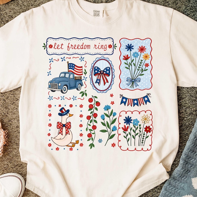 May include: Cream-coloured t-shirt with patriotic graphics. Designs include a blue truck with an American flag, floral arrangements, a bow, a goose in a patriotic hat, and the text "let freedom ring".