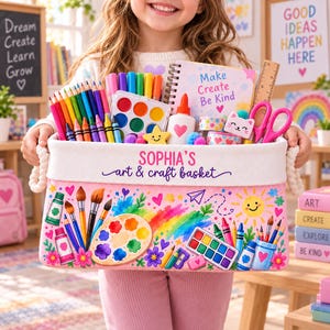 May include: A rectangular fabric storage basket with rope handles, featuring the text 'SOPHIA'S art & craft basket' and colorful illustrations of art supplies. The basket is filled with colored pencils, markers, a watercolor set, a notebook, and scissors.