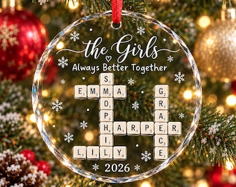 May include: A clear, circular glass Christmas ornament featuring a red ribbon hanger. The design includes white snowflake graphics, the text "the Girls" and "Always Better Together," and a crossword-style arrangement of names: Emma, Sophia, Harper, Lily, and Grace, with the year 2026 at the bottom.
