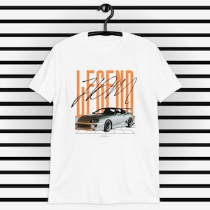 Puede incluir: Camiseta blanca con un gráfico de coche plateado y el texto "LEGEND FROM" en naranja. El coche es un Toyota Supra.