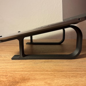 May include: A black laptop stand with a silver laptop resting on top. The stand is made of two curved pieces of metal that form a raised platform.