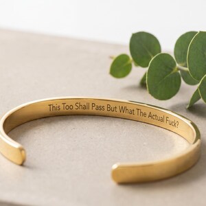 May include: A gold-toned metal cuff bracelet with an open design. The interior surface of the bracelet features the engraved text, "This Too Shall Pass But What The Actual Fuck?" The bracelet has a smooth, polished finish and rounded ends.