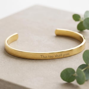 May include: A gold-toned metal cuff bracelet with an open design, featuring the engraved text "This too shall pass" on the outer surface. The bracelet has a smooth, polished finish and a simple, rounded band structure.