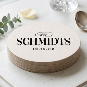 May include: A stack of round, light-colored coasters with a textured surface. The top coaster features black printed text that reads The SCHMIDTS followed by the date 10.15.XX in a clean, sans-serif font centered on the circular surface.