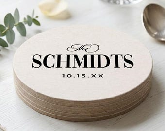 May include: A stack of round, light-colored coasters with a textured surface. The top coaster features black printed text that reads The SCHMIDTS followed by the date 10.15.XX in a clean, sans-serif font centered on the circular surface.
