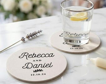 May include: Two round, off-white stone coasters featuring black printed text that reads Rebecca and Daniel with the date 12.18.24 and a small leaf graphic. One coaster sits under a clear glass filled with water and a lemon slice.