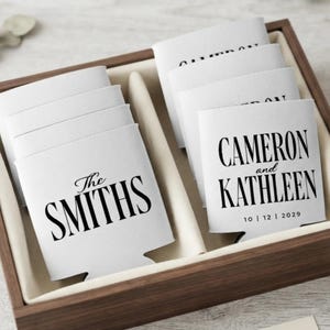 May include: A set of white foam can coolers arranged in a wooden box. The coolers feature black printed text. One reads The SMITHS, while another displays CAMERON and KATHLEEN with the date 10 | 12 | 2029. The sleeves are rectangular with a cutout base.