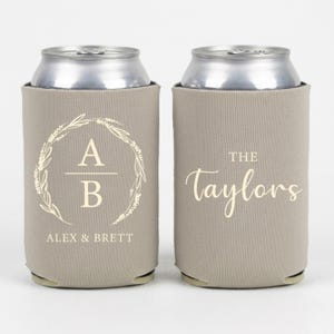 May include: Two light beige foam can coolers. The left cooler features a cream-colored botanical wreath surrounding the letters A and B with the text ALEX & BRETT below. The right cooler displays the text THE Taylors in cream lettering.