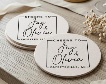 May include: Two round, off-white coasters featuring a black printed design. The text reads "CHEERS TO" above "Jay & Olivia" inside a square border, with "FAYETTEVILLE, AK" printed below. The coasters have a smooth, matte finish.