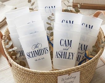 May include: A woven basket filled with frosted plastic cups featuring dark blue printed text. The cups display names like CAM and ASHLEY, or THE SCHMIDTS with the date 10.15.XX. A white tag attached to the basket front reads Wedding Favors.