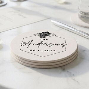 May include: A stack of three round, light beige coasters featuring a black hexagonal border, a floral illustration at the top, and the text "THE Andersons" followed by the date "06.11.2024" printed in the center of each coaster.