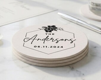 May include: A stack of three round, light beige coasters featuring a black hexagonal border, a floral illustration at the top, and the text "THE Andersons" followed by the date "06.11.2024" printed in the center of each coaster.
