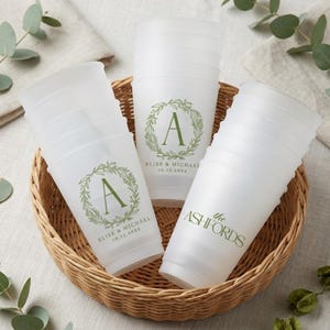 May include: A set of translucent white plastic cups stacked in a woven basket. The cups feature green printed designs, including a letter A inside a botanical wreath with the text Elise & Michael 10.15.20XX and the text the ASHFORDS.