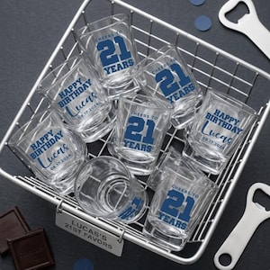 May include: A collection of clear glass shot glasses arranged in a wire basket. Some glasses feature the blue text 'CHEERS TO 21 YEARS', while others display 'HAPPY BIRTHDAY LUCAS 08.17.2024'. A metal tag attached to the basket reads 'LUCAS'S 21ST FAVORS'.
