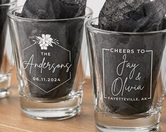 Puede incluir: Dos vasos de chupito transparentes rellenos de papel de seda negro. El vaso izquierdo presenta un marco hexagonal blanco con un diseño floral, el texto The Andersons y 06.11.2024. El vaso derecho tiene un marco cuadrado blanco, el texto Cheers to Jay & Olivia y Fayetteville, AK.