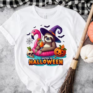 May include: White t-shirt with a Halloween-themed design. It features a sloth in a witch's hat on a pink flamingo float, holding a drink. A jack-o'-lantern, skull, and the text "WAITING ON HALLOWEEN" are also included.