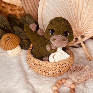 May include: A handmade green crochet dinosaur sits in a woven basket, holding a white cloth. A wooden teething ring and a tassel are nearby. The background includes a decorative fan and a mushroom-shaped object, all on a white, fluffy surface.
