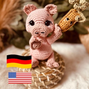 Könnte beinhalten: Ein rosa gehäkeltes Schwein, das eine braune gehäkelte Karotte hält. Das Schwein hat schwarze Augen und eine rosa Nase. Die Karotte hat ein braunes Gesicht mit schwarzen Augen. Das Schwein und die Karotte sitzen auf einem geflochtenen Korb.