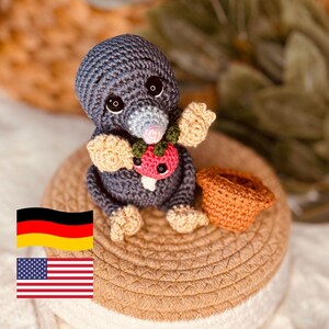 Può includere: Una bambola di talpa all'uncinetto fatta a mano nei toni del grigio, beige e rosa, che tiene in mano una fragola rossa. La talpa è seduta su un cestino intrecciato con un cestino all'uncinetto arancione nelle vicinanze. La bambola ha grandi occhi neri.