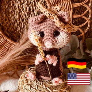 Op de afbeelding: Een gehaakte hamster die een strohoed draagt en een tarwehalm vasthoudt. De hamster zit op een geweven mand met een Duitse vlag en een Amerikaanse vlag op de achtergrond.