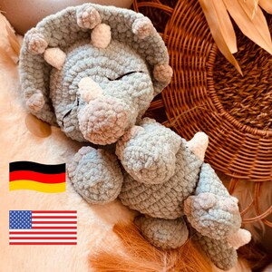 May include: A crocheted plush toy in the shape of a triceratops dinosaur. The toy is light blue with white accents and has a sleepy expression. The toy is lying on a brown woven surface.
