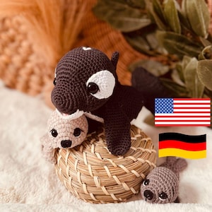 Puede incluir: Tres animales de peluche tejidos a crochet: una orca negra y blanca, una foca marrón claro y una foca marrón oscuro. La orca está sentada en una cesta tejida con las dos focas delante de ella. El fondo es una imagen borrosa de una planta marrón y verde.