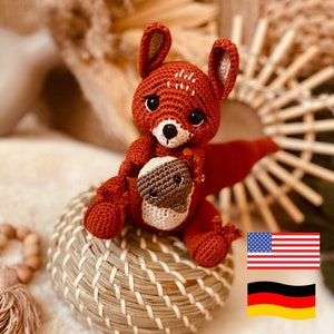May include: A crocheted fox amigurumi toy holding a smaller crocheted animal. The fox is orange and brown with black eyes and a white nose. The smaller animal is brown and white with black eyes. The toys are sitting on a woven basket.