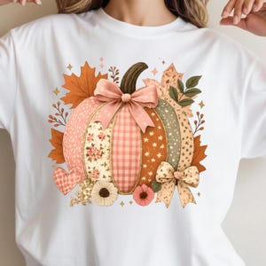 May include: White t-shirt featuring a colorful pumpkin design. The pumpkin is segmented with various patterns, including floral, gingham, and dotted sections. It is adorned with a pink bow, autumn leaves, and floral accents, creating a fall-themed graphic.