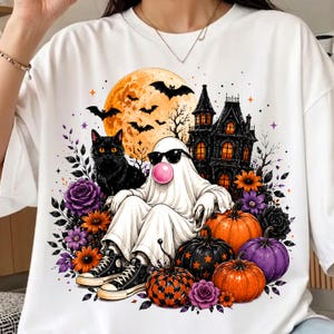 May include: A white t-shirt featuring a graphic print of a ghost wearing sunglasses and blowing a pink bubble, seated among pumpkins, a black cat, a haunted house, bats, a full moon, and purple and orange flowers with black sneakers.