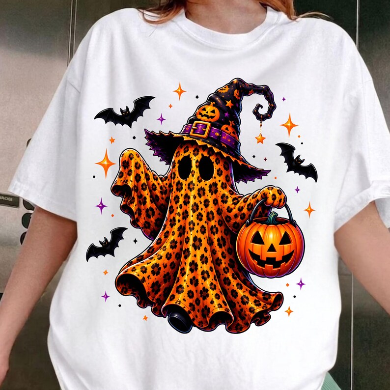 May include: A white short-sleeve t-shirt featuring a graphic of a ghost with an orange and black leopard print pattern. The ghost wears a matching witch hat with a pumpkin and holds an orange jack-o'-lantern bucket, surrounded by black bats.