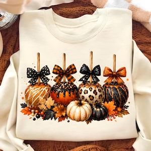 May include: Cream-colored sweatshirt featuring a festive fall design. The graphic showcases four caramel apples with decorative bows, surrounded by autumn leaves and pumpkins. The apples have various patterns and colors.